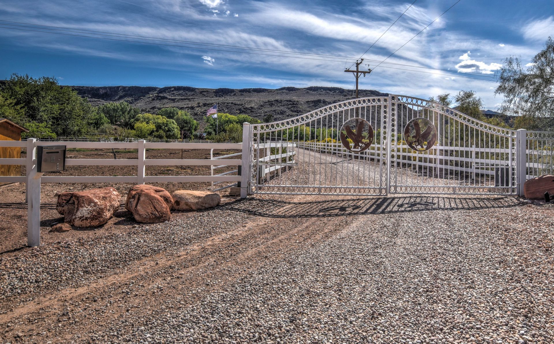 White gated entrance and wooden fence beside a gravel road in a desert landscape under a blue sky