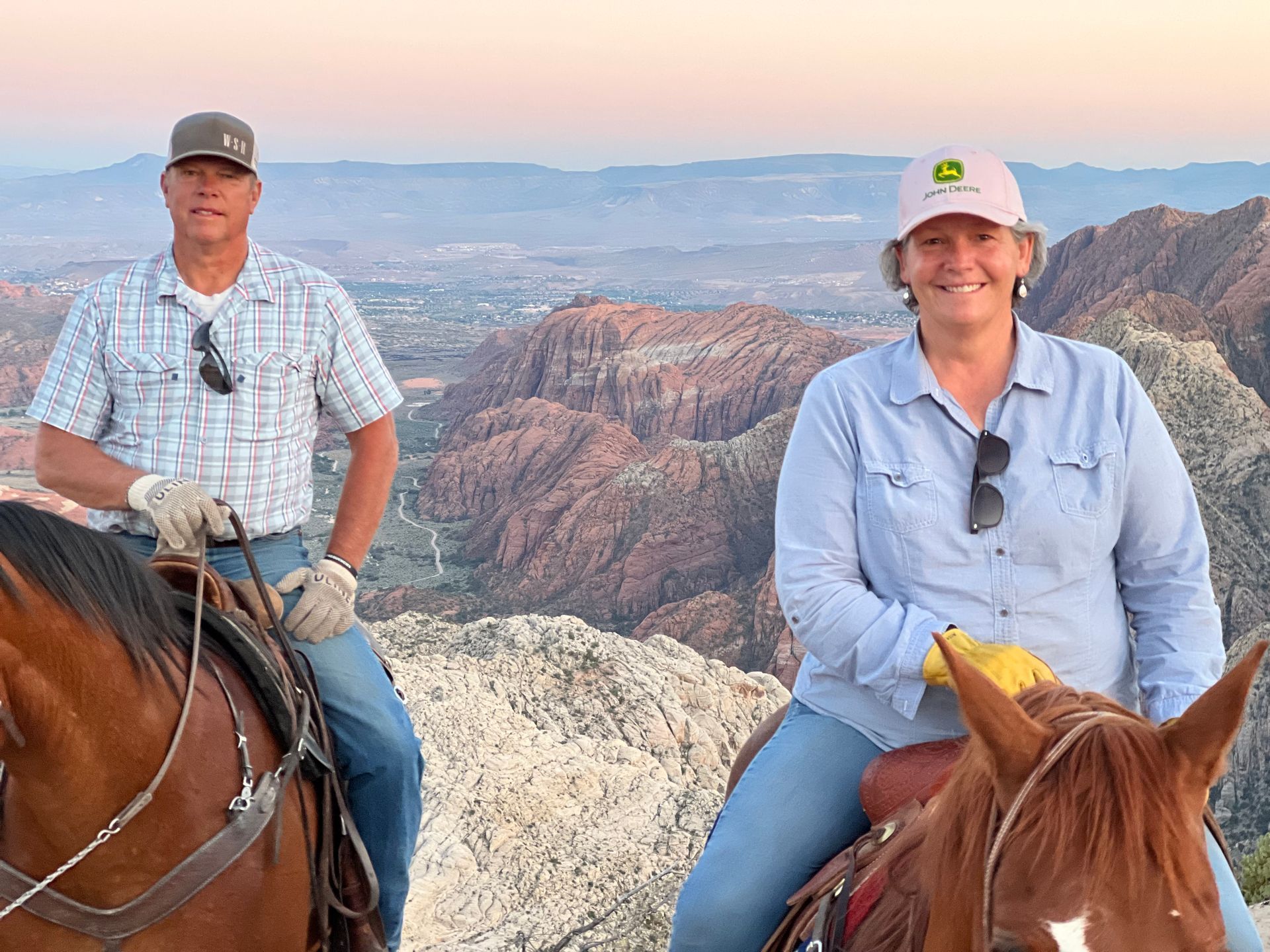 Two horseback riders in helmets on a canyon overlook at sunset