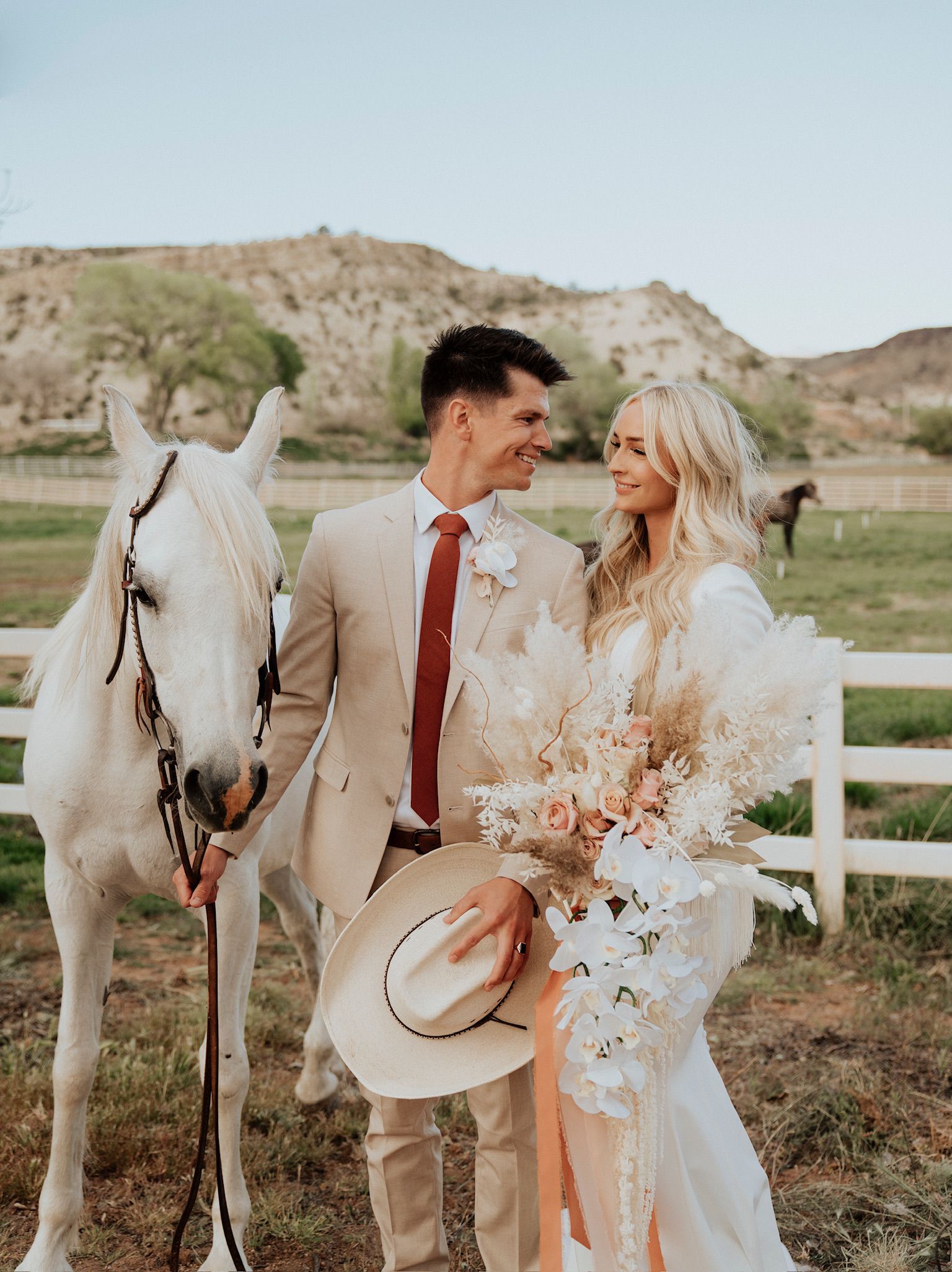 Couple posing with a white horse in a grassy field, wearing cream wedding attire, with hills and a fence behind them