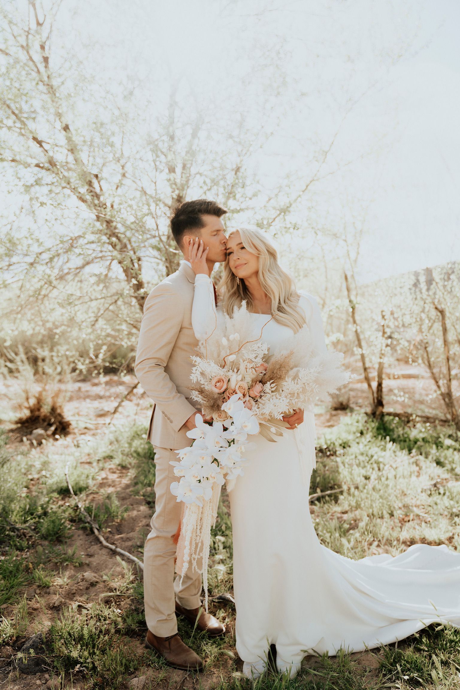 Bride and groom embracing in a sunlit orchard, holding a bouquet of white flowers, with a long wedding dress.