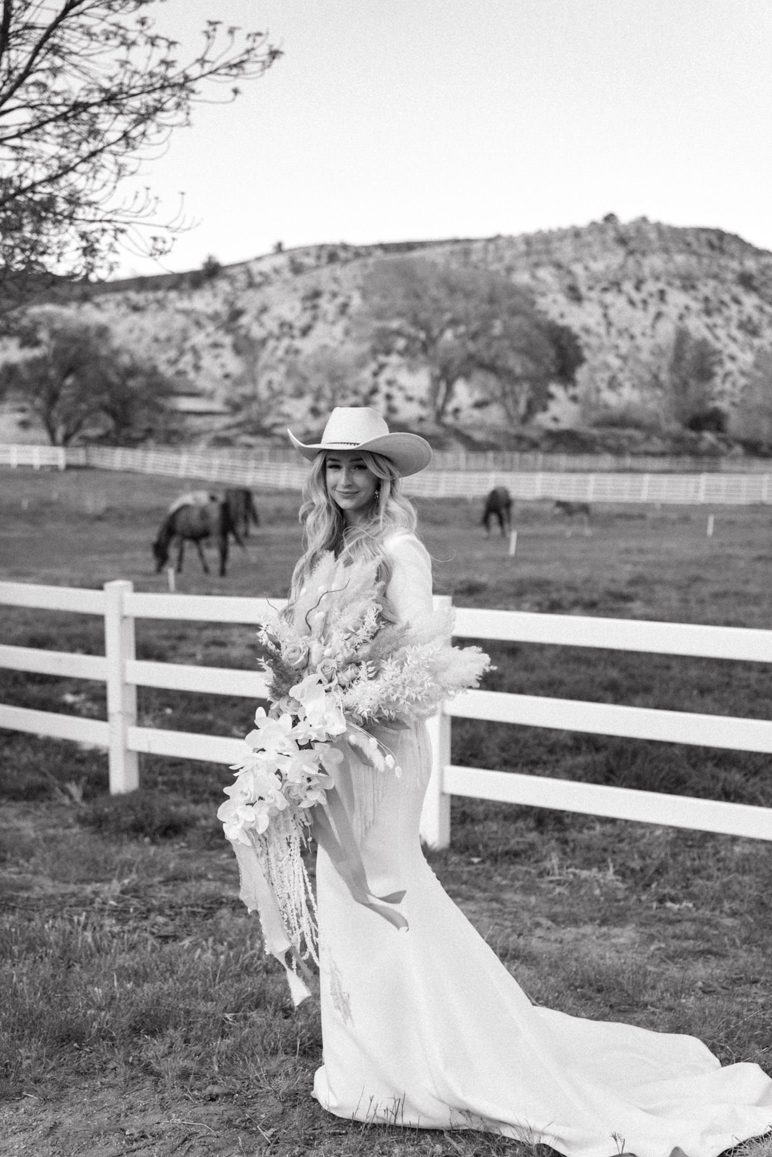 Woman in a long white dress and cowboy hat holding flowers beside a white fence in a pasture