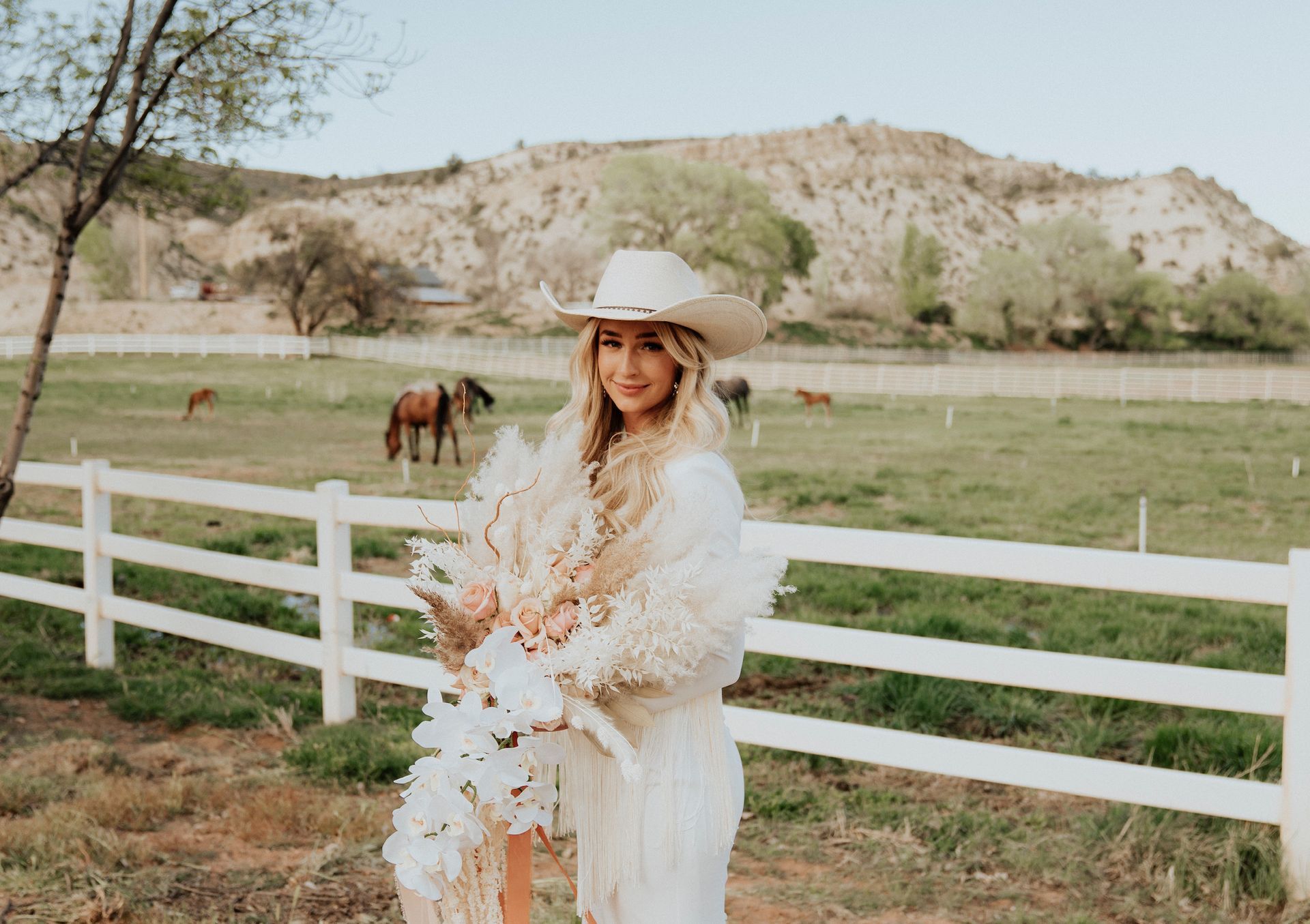 Woman in a white dress and cowboy hat holding flowers by a white fence with horses and hills behind her