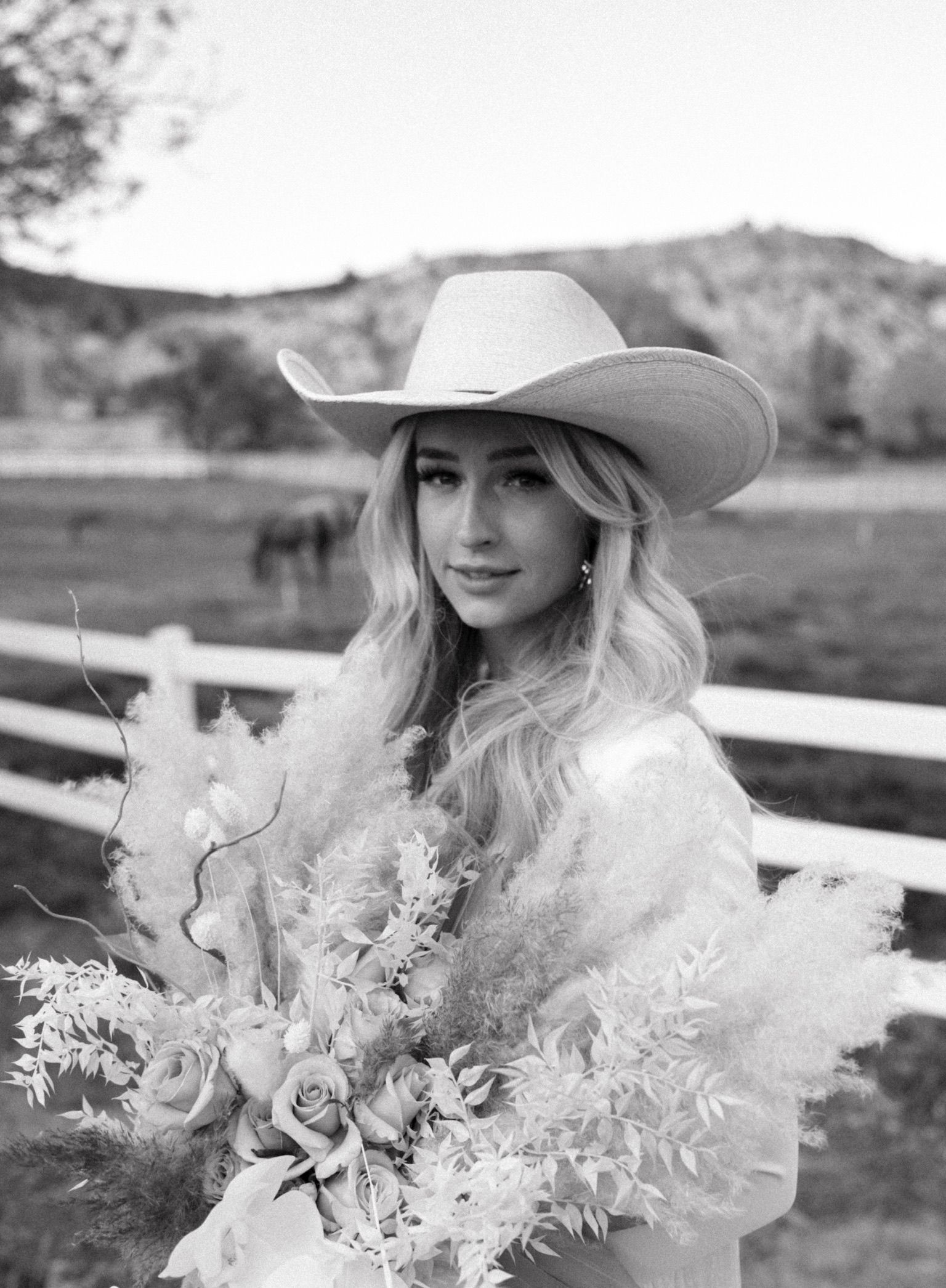 Woman in a cowboy hat holding flowers by a fence in a rural field