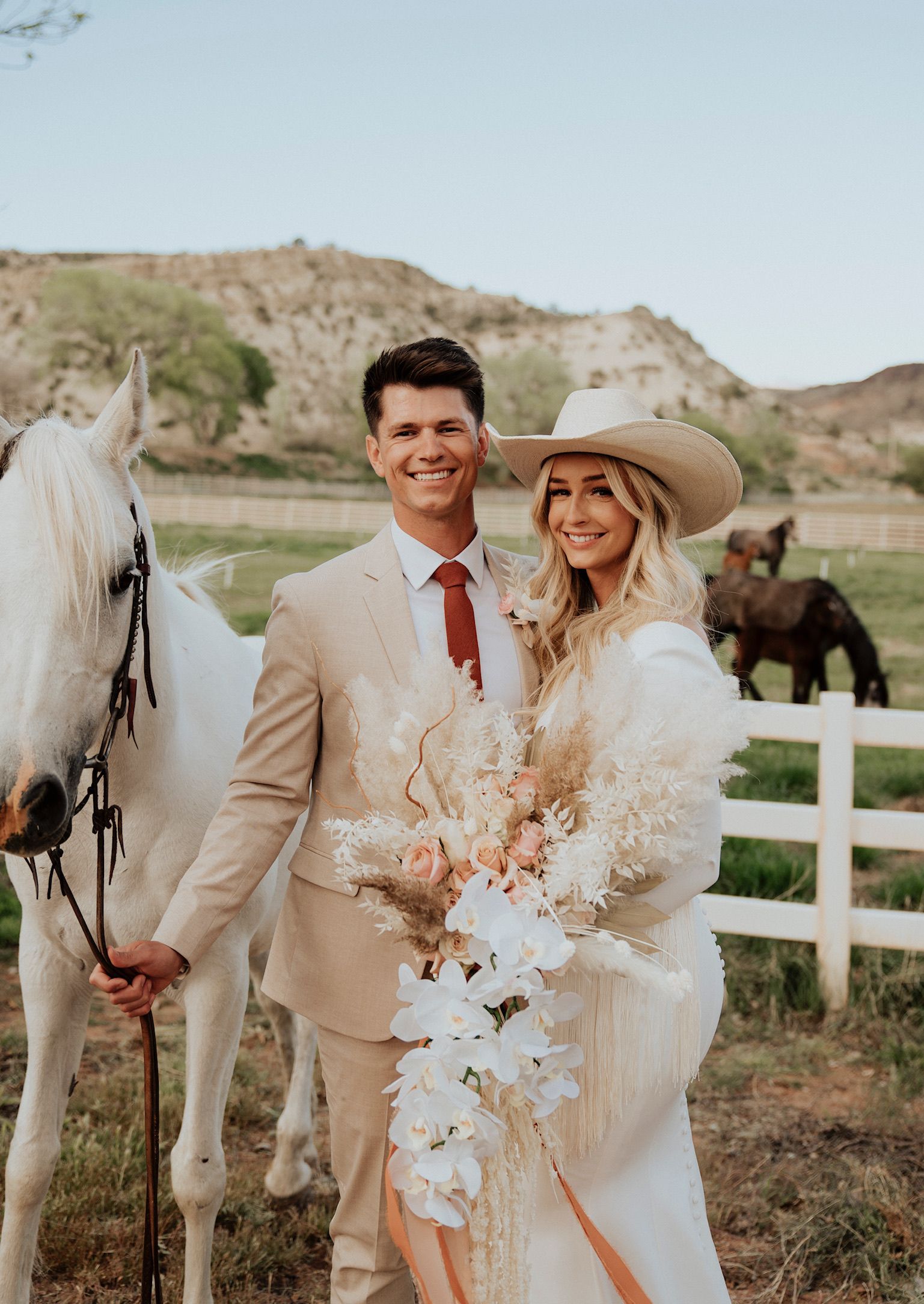 Wedding couple in beige outfits standing with a white horse in a rustic field