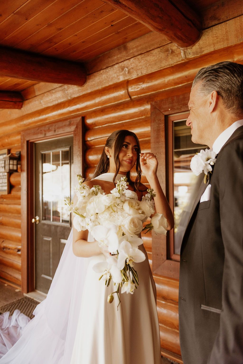 Bride in white gown holding bouquet on a wooden porch, facing a groom in a tuxedo