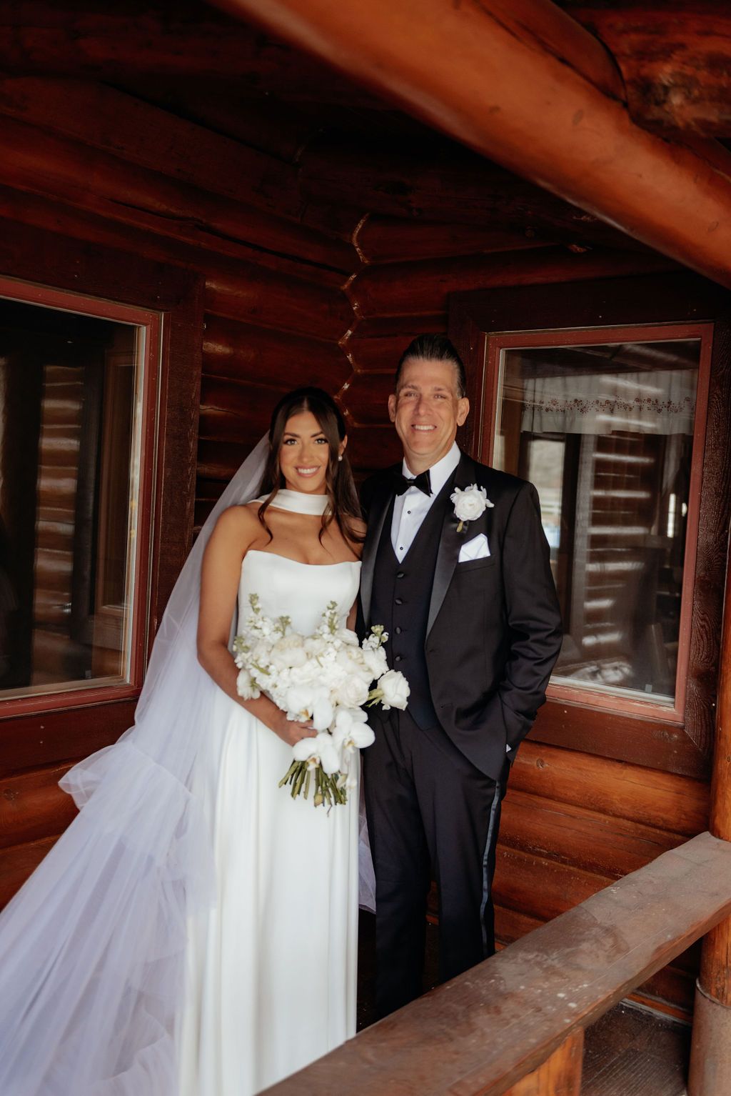 Bride and groom posing on a wooden balcony, bride holding a white bouquet and groom in a black tuxedo.