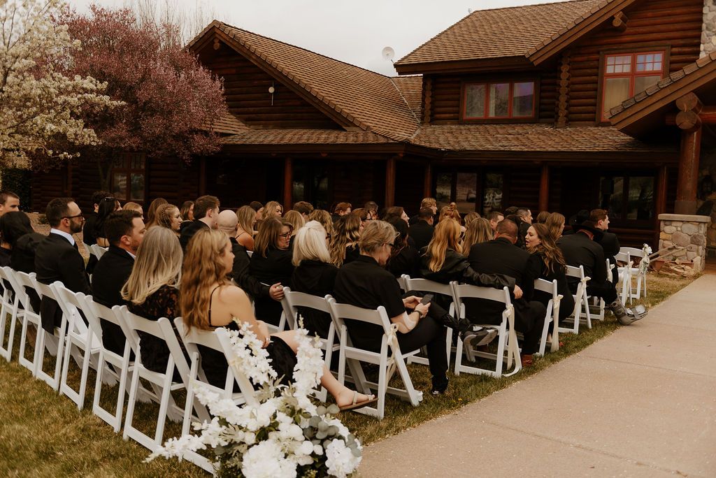 Outdoor wedding ceremony with guests seated in white chairs beside a rustic wooden lodge