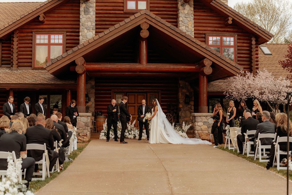 Bride and groom at outdoor wedding ceremony in front of a rustic lodge, guests seated on both sides