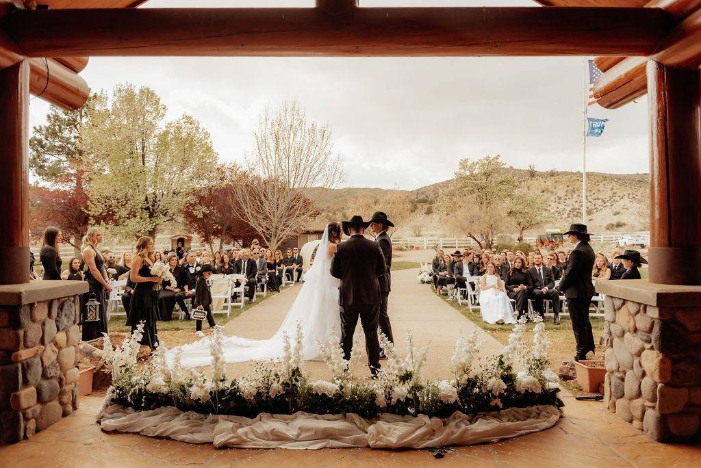 Outdoor wedding ceremony under a rustic pavilion, with bride and groom at the altar and seated guests on both sides