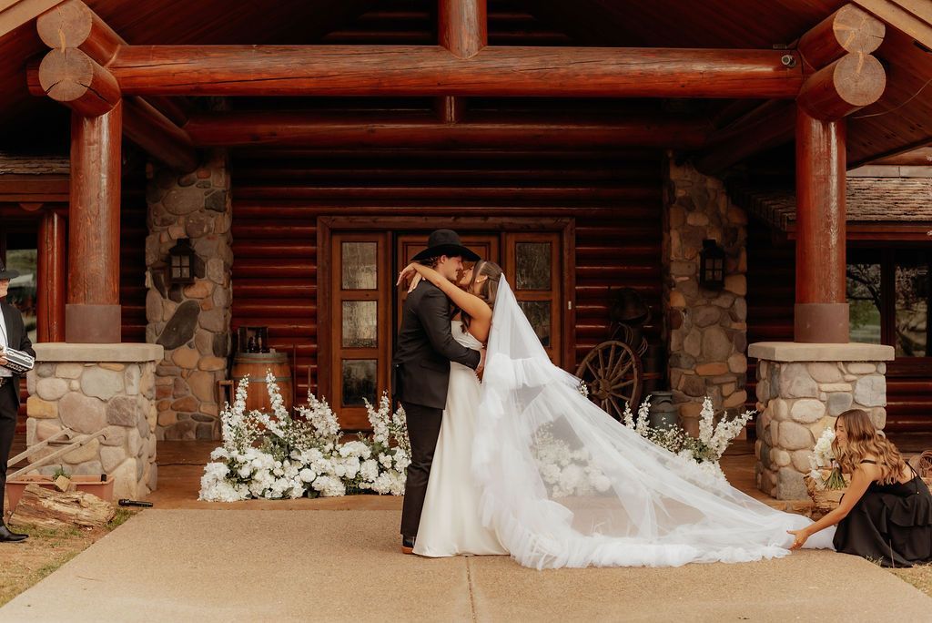 Bride and groom kiss in front of a rustic lodge with white floral decor and a long veil flowing behind her