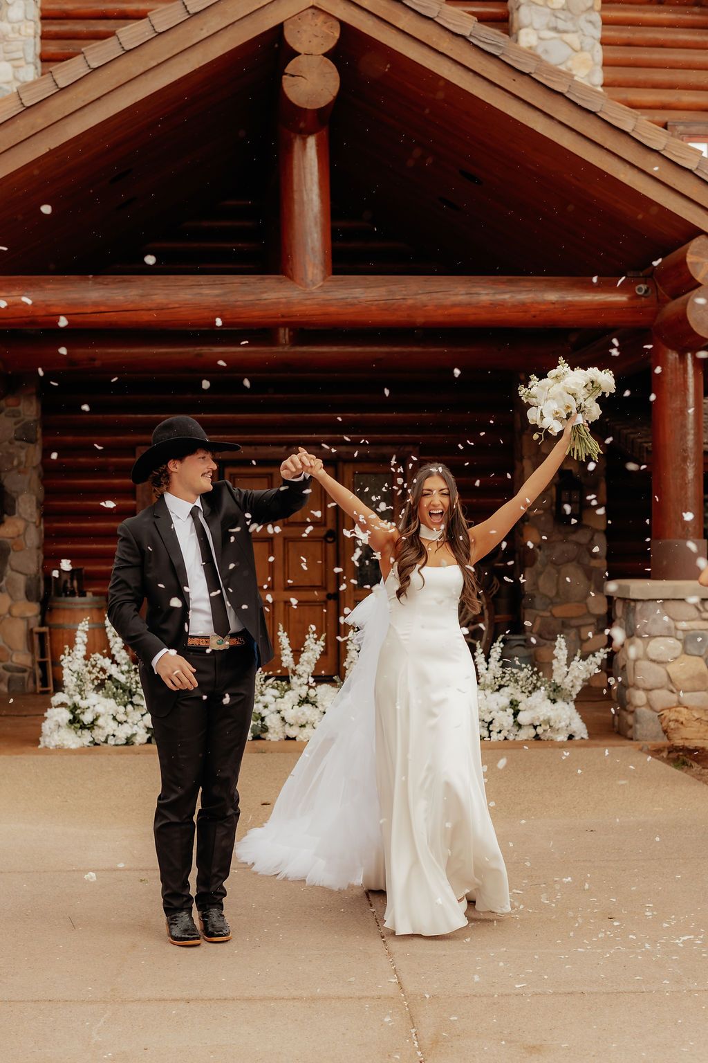 Couple in wedding attire outside a rustic cabin, raising hands amid falling snow.