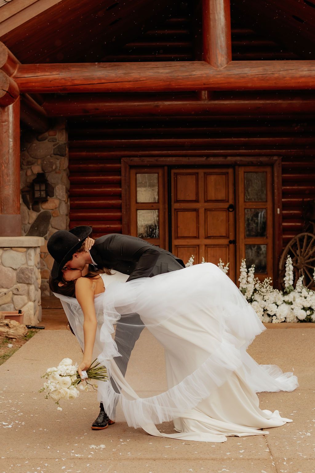Wedding couple kissing in front of a rustic cabin, bride in white dress and groom in black suit