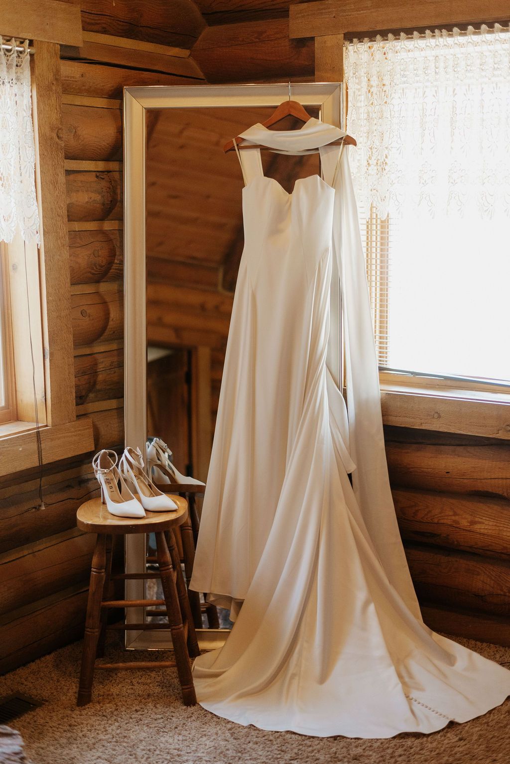 Wedding dress hanging in a rustic cabin by a window, with white shoes on a stool nearby