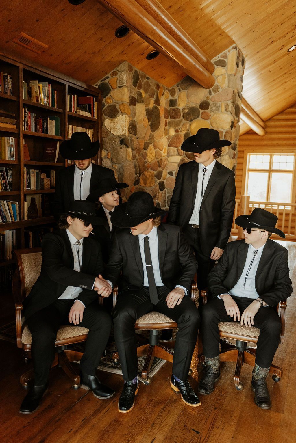 Five men in black suits and hats pose in a rustic cabin with a stone fireplace and wood ceiling.