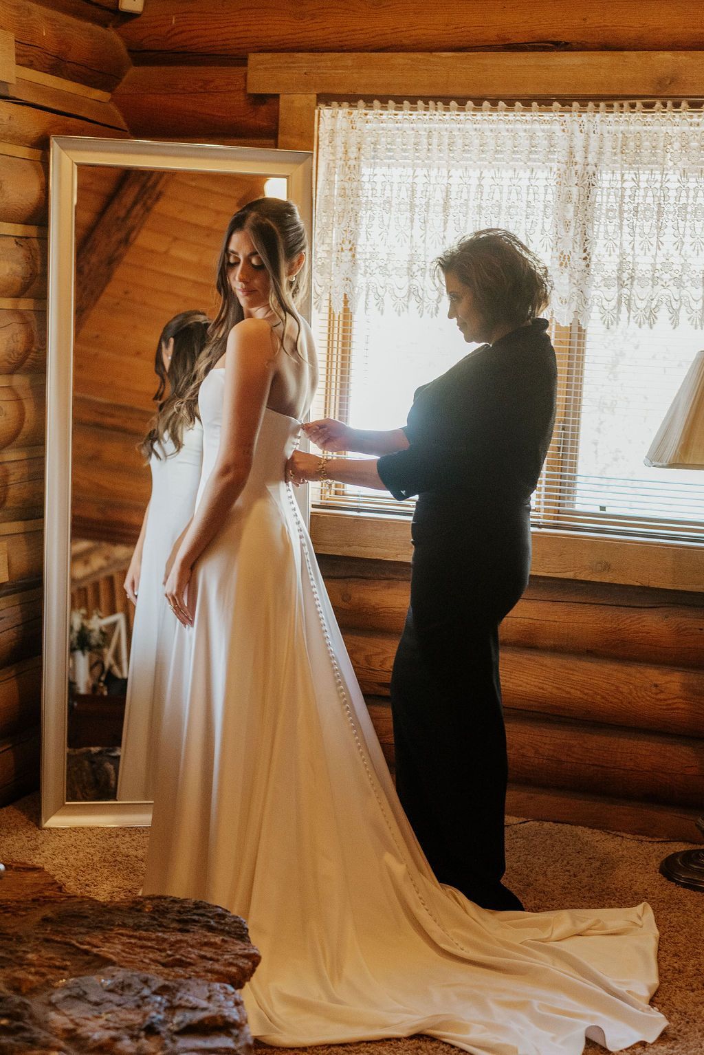 Bride in white dress getting ready with a stylist in a rustic log cabin by a lace-curtained window