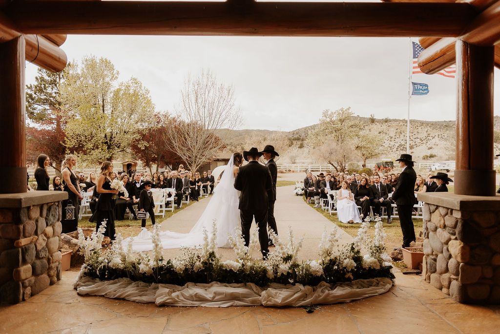 Wedding ceremony under a wooden pavilion, with bride and groom standing on a flower-lined aisle.