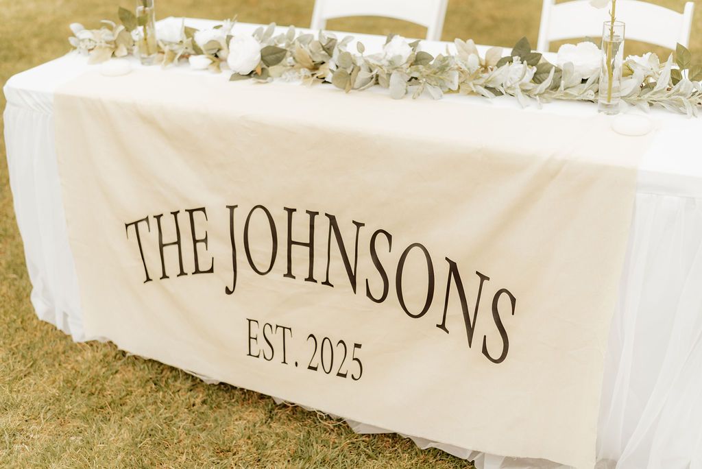 White table with floral centerpiece and “THE JOHNSONS EST. 2026” on the front at an outdoor event