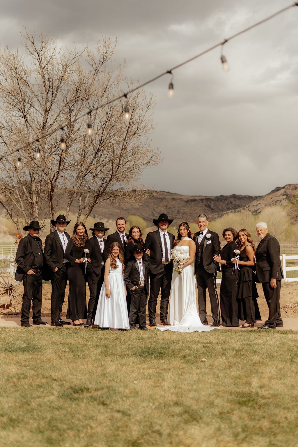 Wedding party posing outdoors with string lights, mountains, and two brides in white dresses at center