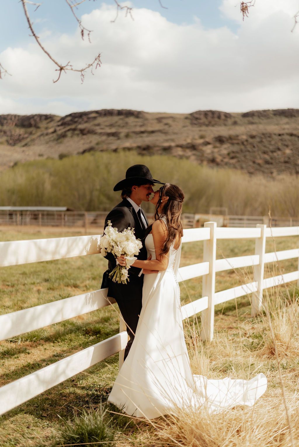 Couple in wedding attire embracing by a white fence in a grassy field under a cloudy sky