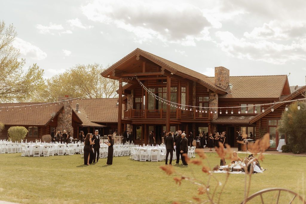 Outdoor wedding reception at a rustic wooden lodge with guests seated on a lawn and a decorated arch nearby