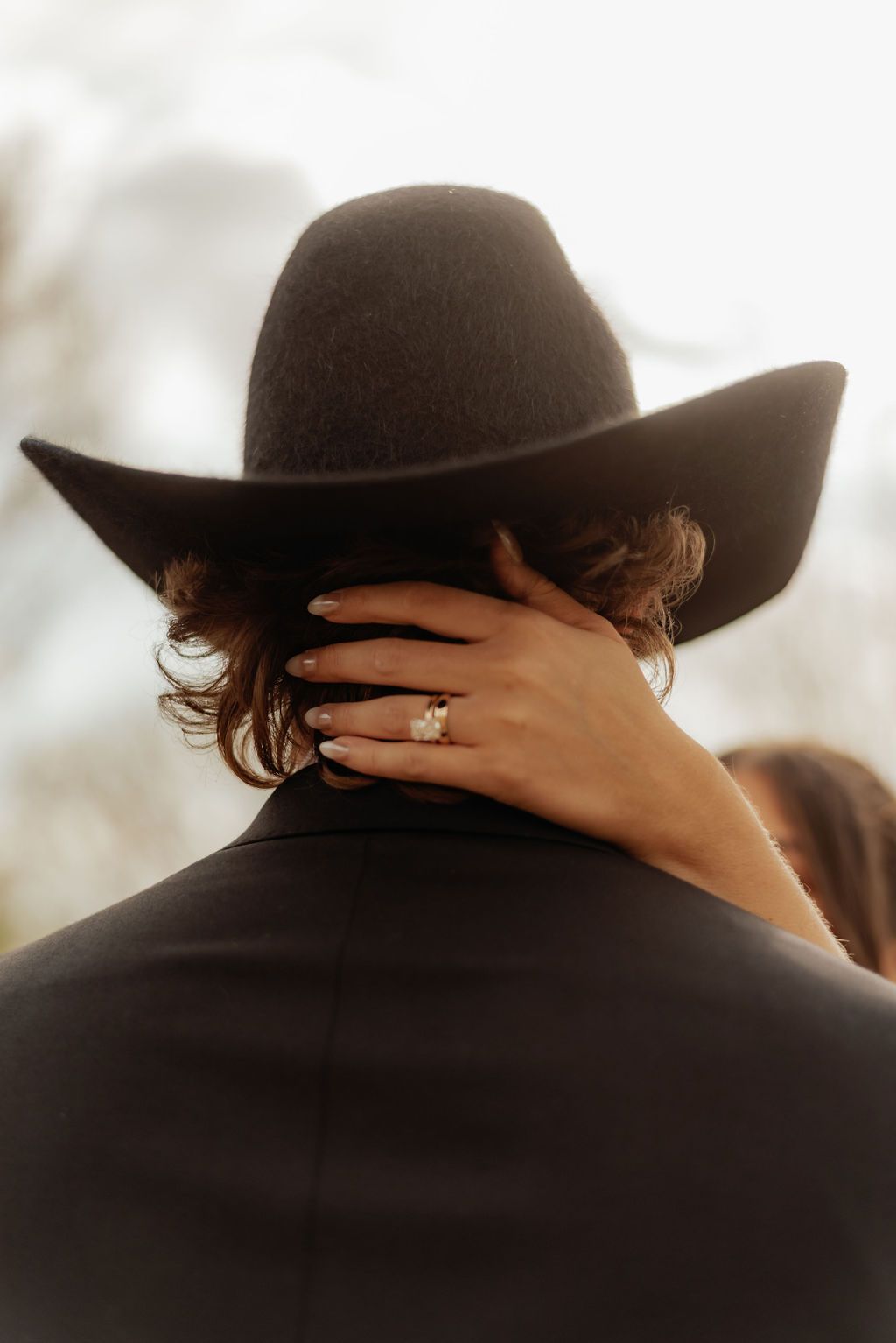 Hand in ring rests on shoulder beneath a black cowboy hat, with curly hair and soft warm light.