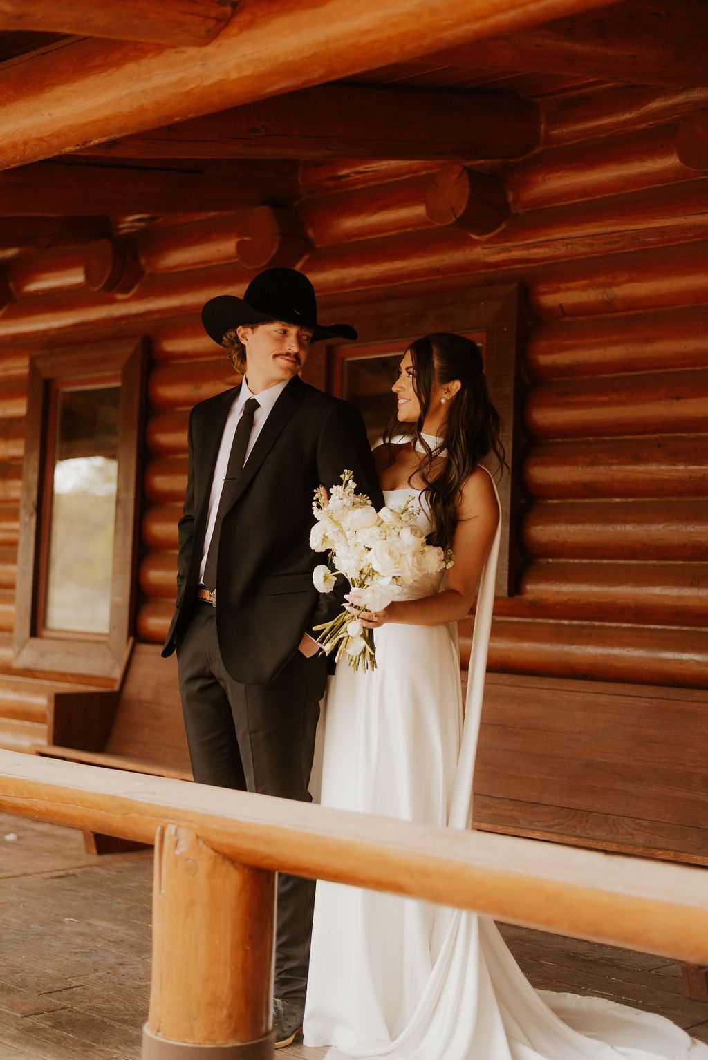 Couple in wedding attire standing on a wooden porch with log cabin walls