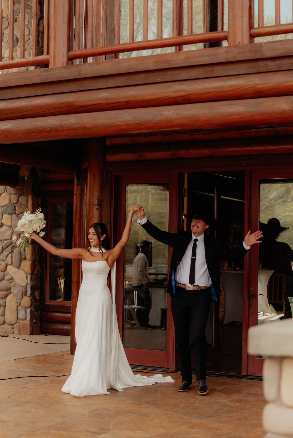 Bride and groom in white and black standing in a wooden doorway with arms raised