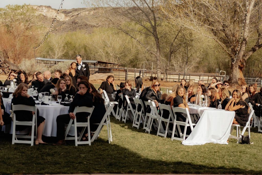 Guests seated at an outdoor event on a grassy lawn with white chairs and trees in the background