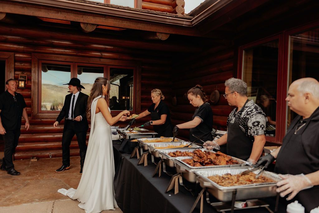Bride and guests at a buffet table in a wooden lodge, serving food during a wedding reception