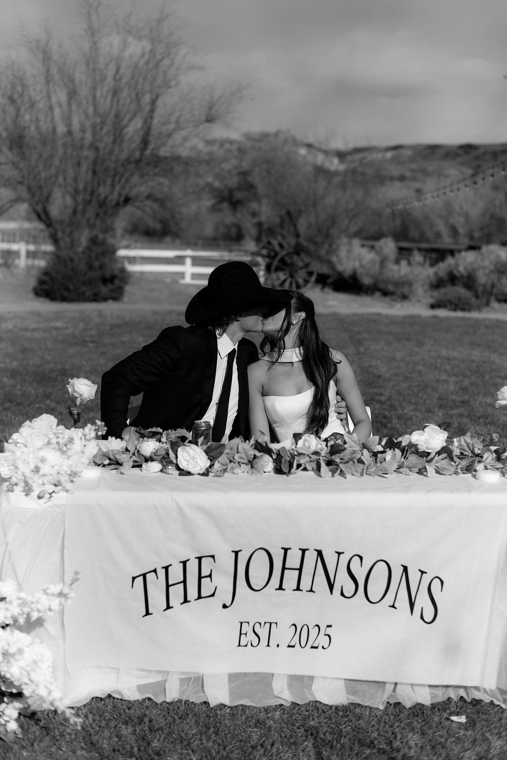 Couple leaning together at a decorated table with “THE JOHNSTONS EST. 2025” sign outdoors.