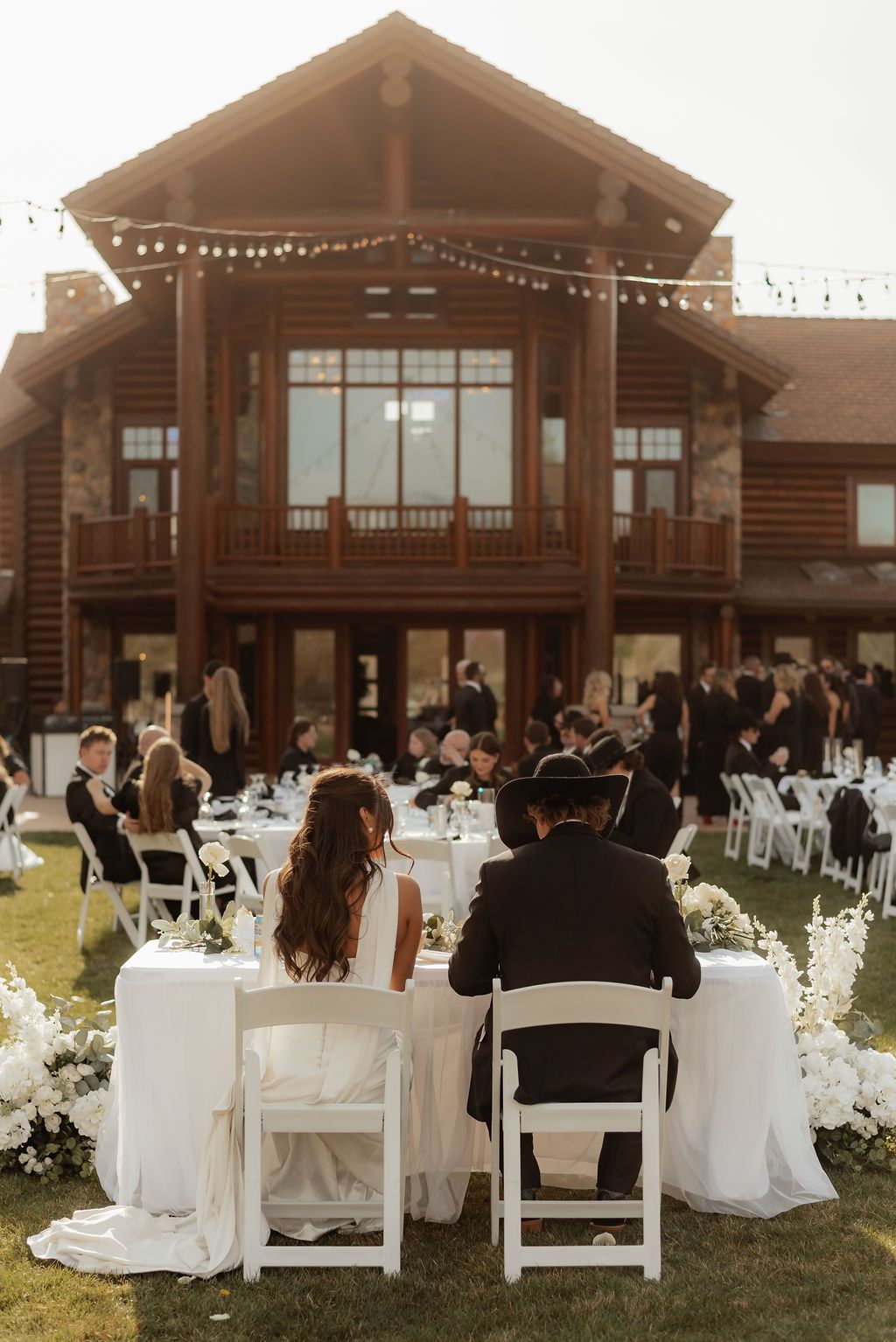 Wedding ceremony in front of a rustic wooden lodge, with the bride and groom seated facing guests outdoors.
