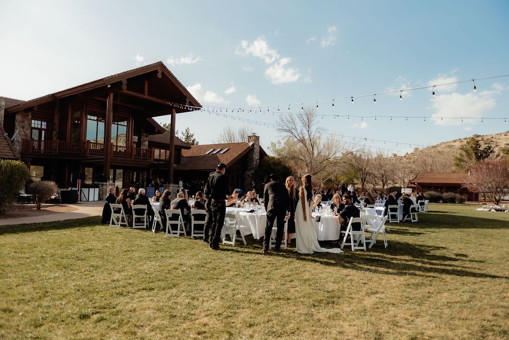 Outdoor wedding ceremony with guests seated on a lawn beside a wooden lodge under a clear sky