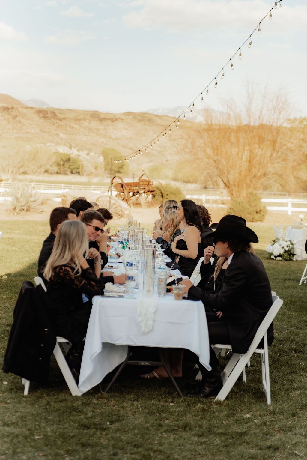 Outdoor banquet table with guests seated in black attire under string lights, overlooking a sandy landscape