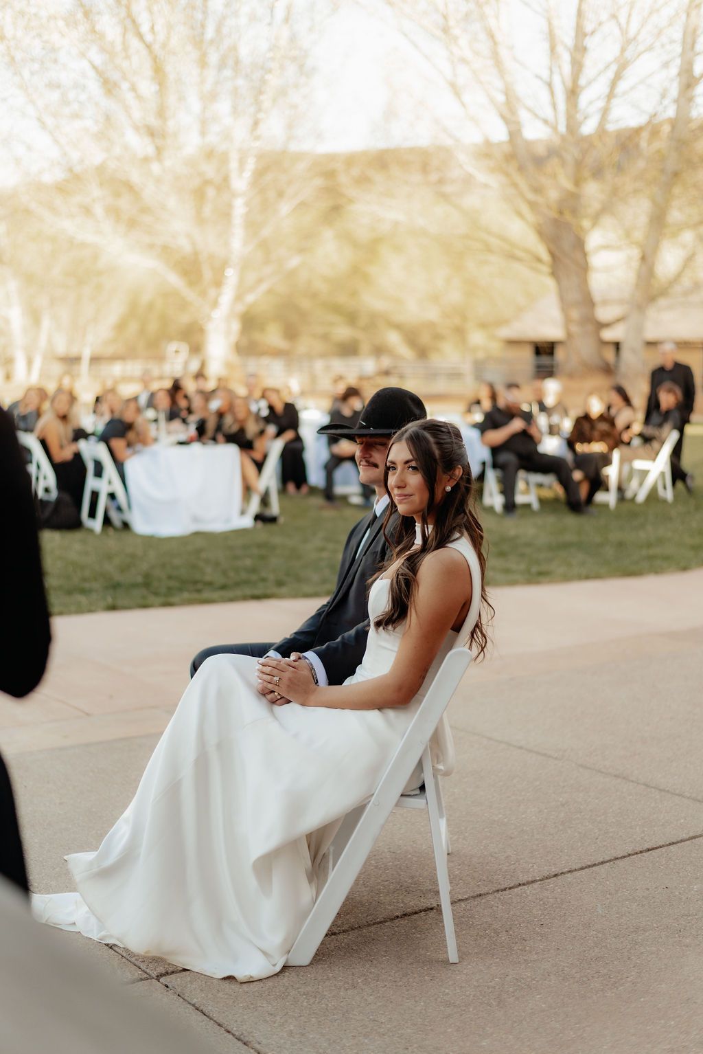 Woman in a white dress sitting on a chair at an outdoor reception with guests in the background