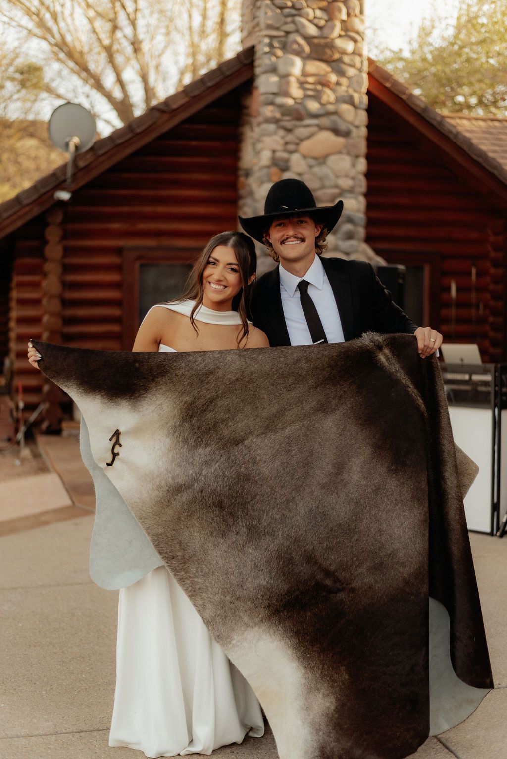 Couple holding a large brown-and-white hide in front of a log cabin and stone chimney