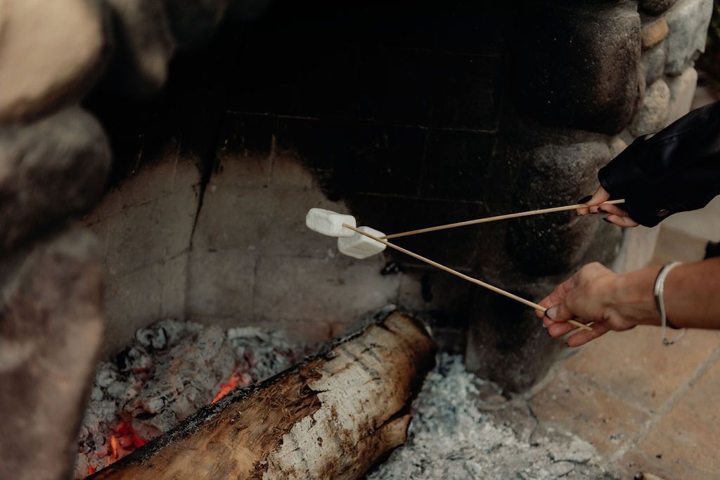 Hands roasting marshmallows over a campfire with embers and logs in a stone hearth