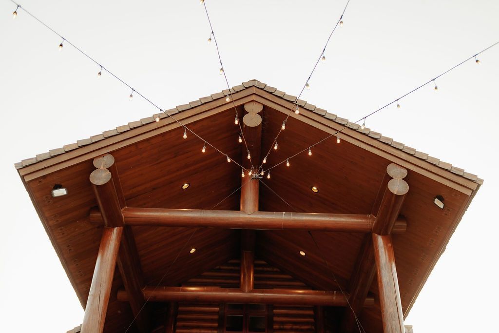 Low-angle view of a wooden pavilion roof with exposed beams and hanging string lights against the sky