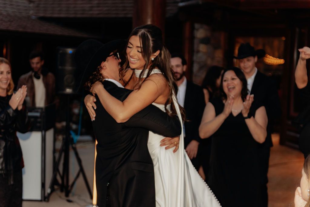 Two women in black and white dresses hug at a lively indoor celebration with guests applauding nearby