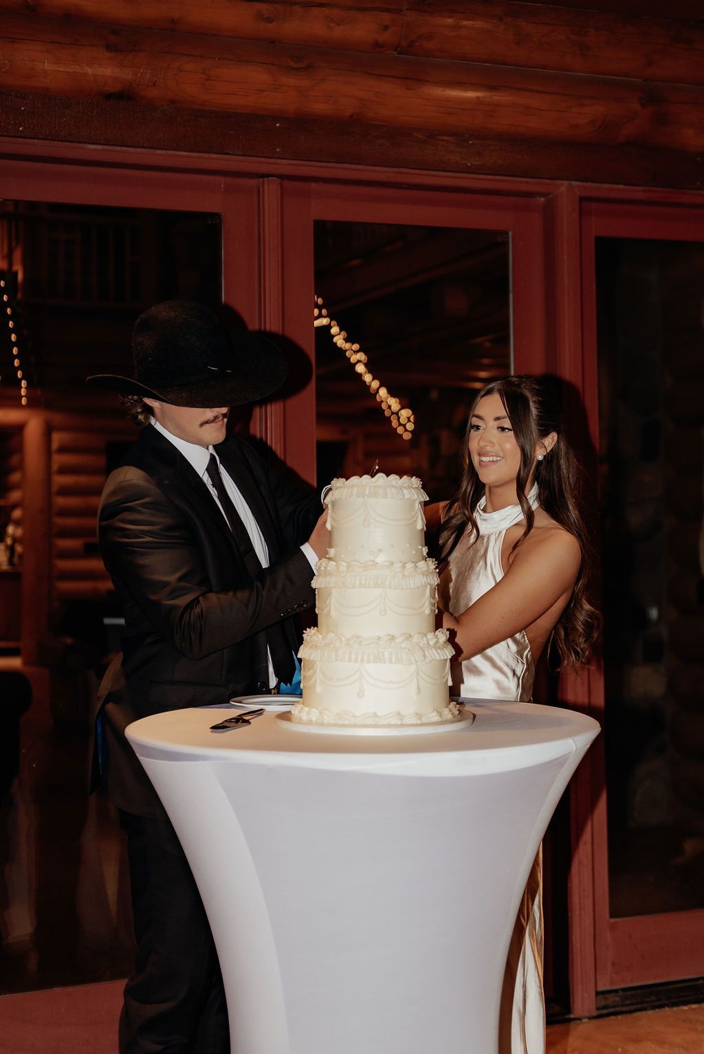 Couple cutting a tiered wedding cake beside a tall cocktail table in a dimly lit reception hall