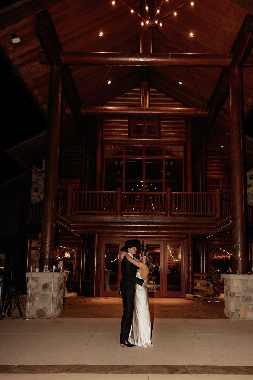 Couple embracing in formal attire before a warmly lit wooden lodge at night