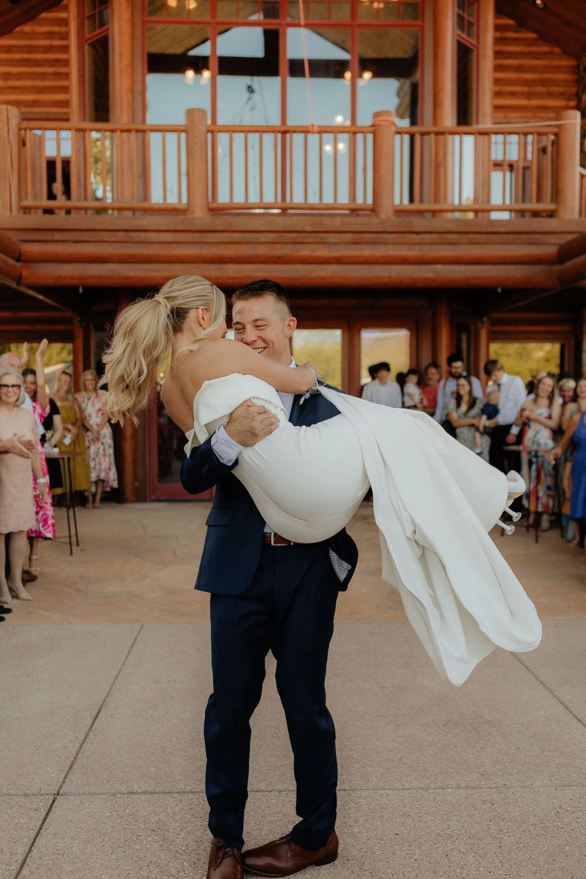 Wedding couple embracing outside a log cabin, with guests gathered in the background