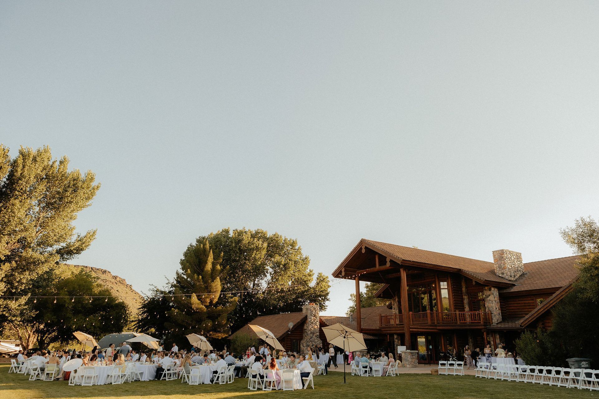 Outdoor wedding reception with white chairs and guests on a lawn beside a wooden lodge at sunset