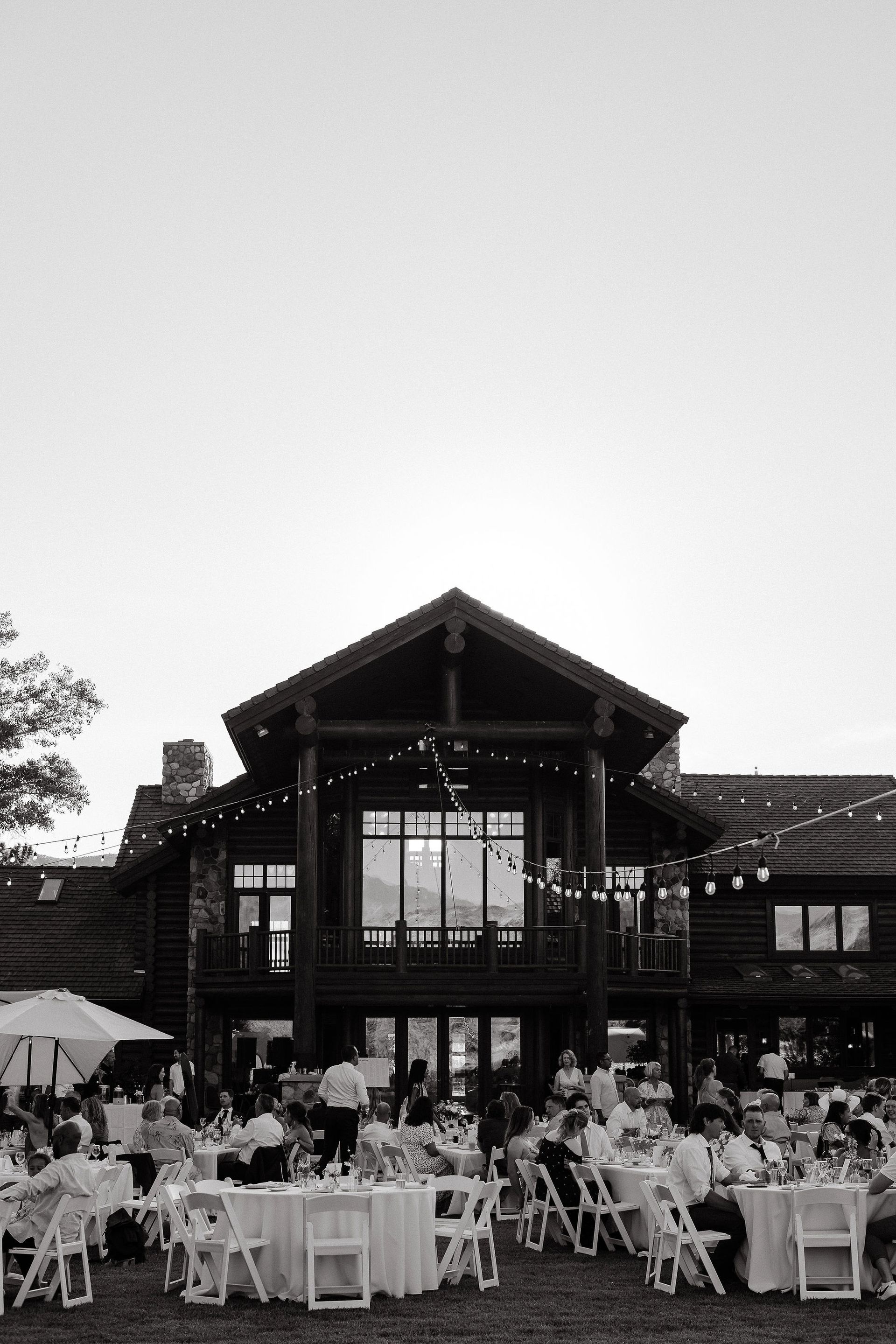 Outdoor reception with guests seated at tables in front of a large rustic lodge at dusk