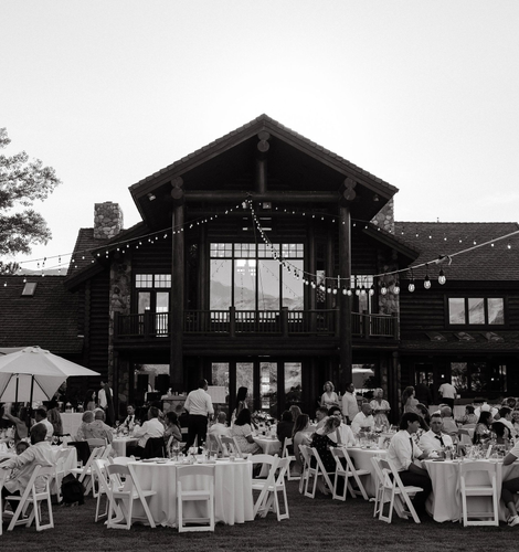 Outdoor wedding reception under a large wooden lodge, with guests seated at white tables.