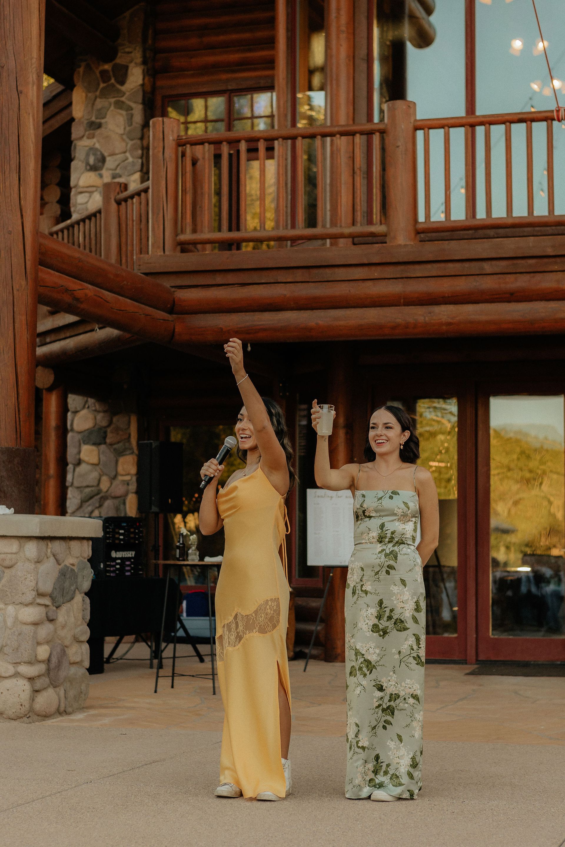 Two women in formal dresses standing outside a rustic lodge, one raising an arm and the other holding a sign.