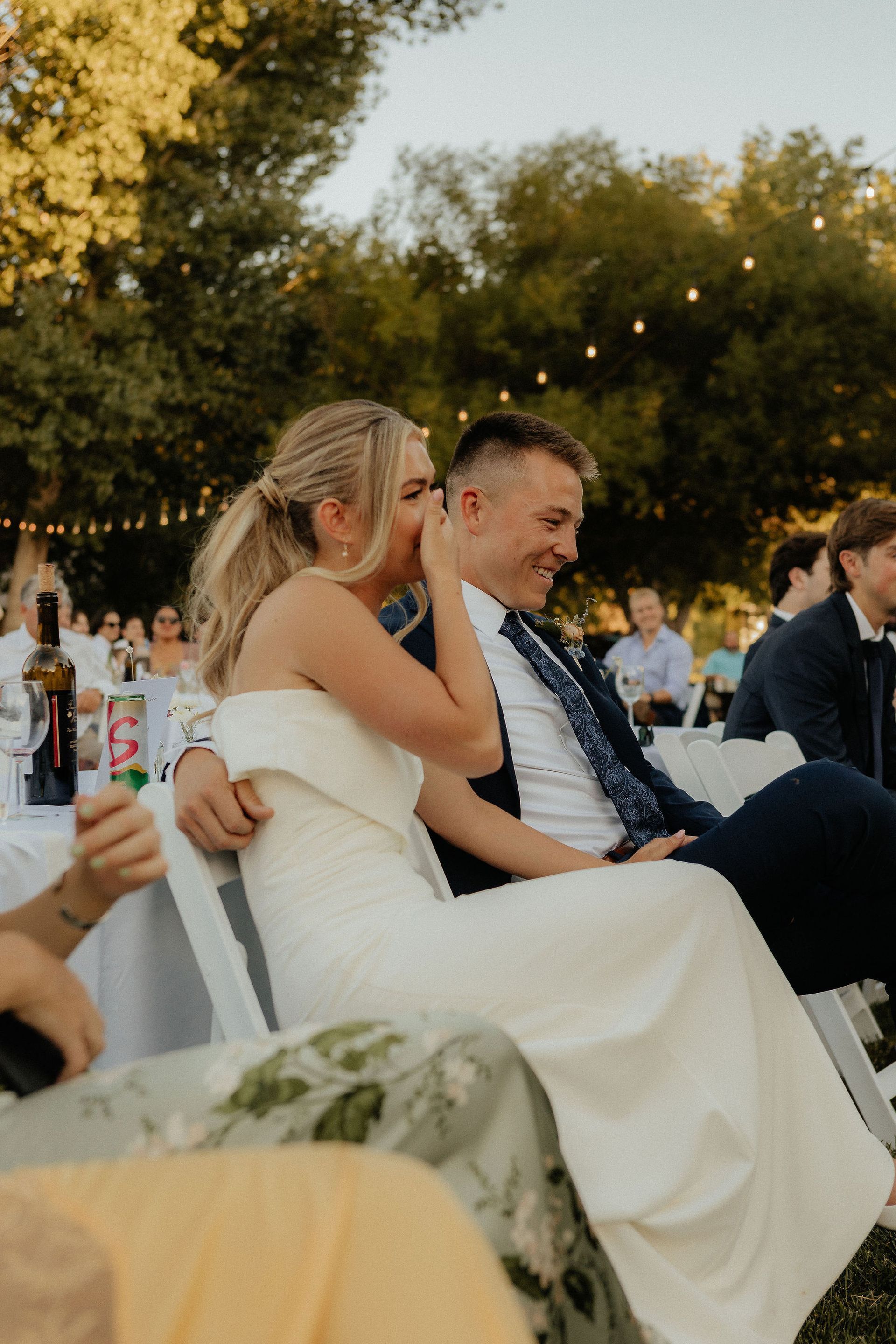 Couple seated at an outdoor evening celebration, smiling among guests and string lights