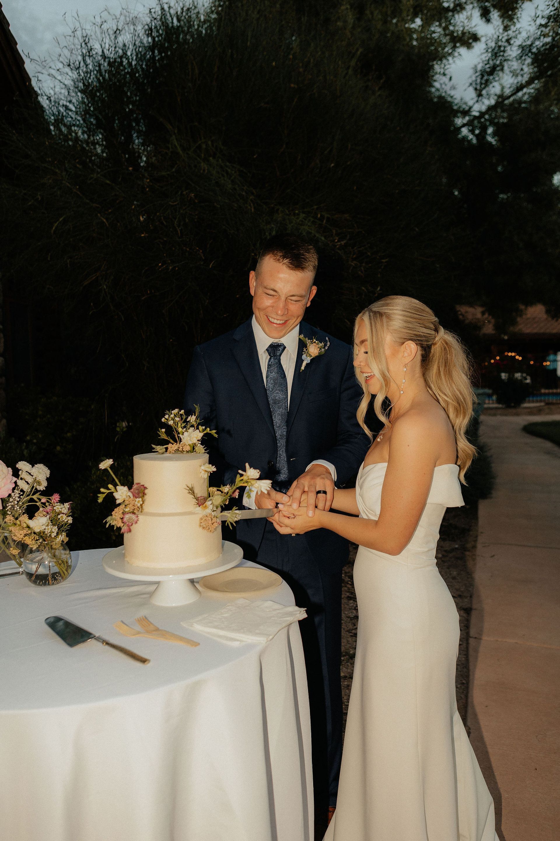Bride and groom cutting a wedding cake at an outdoor reception table.
