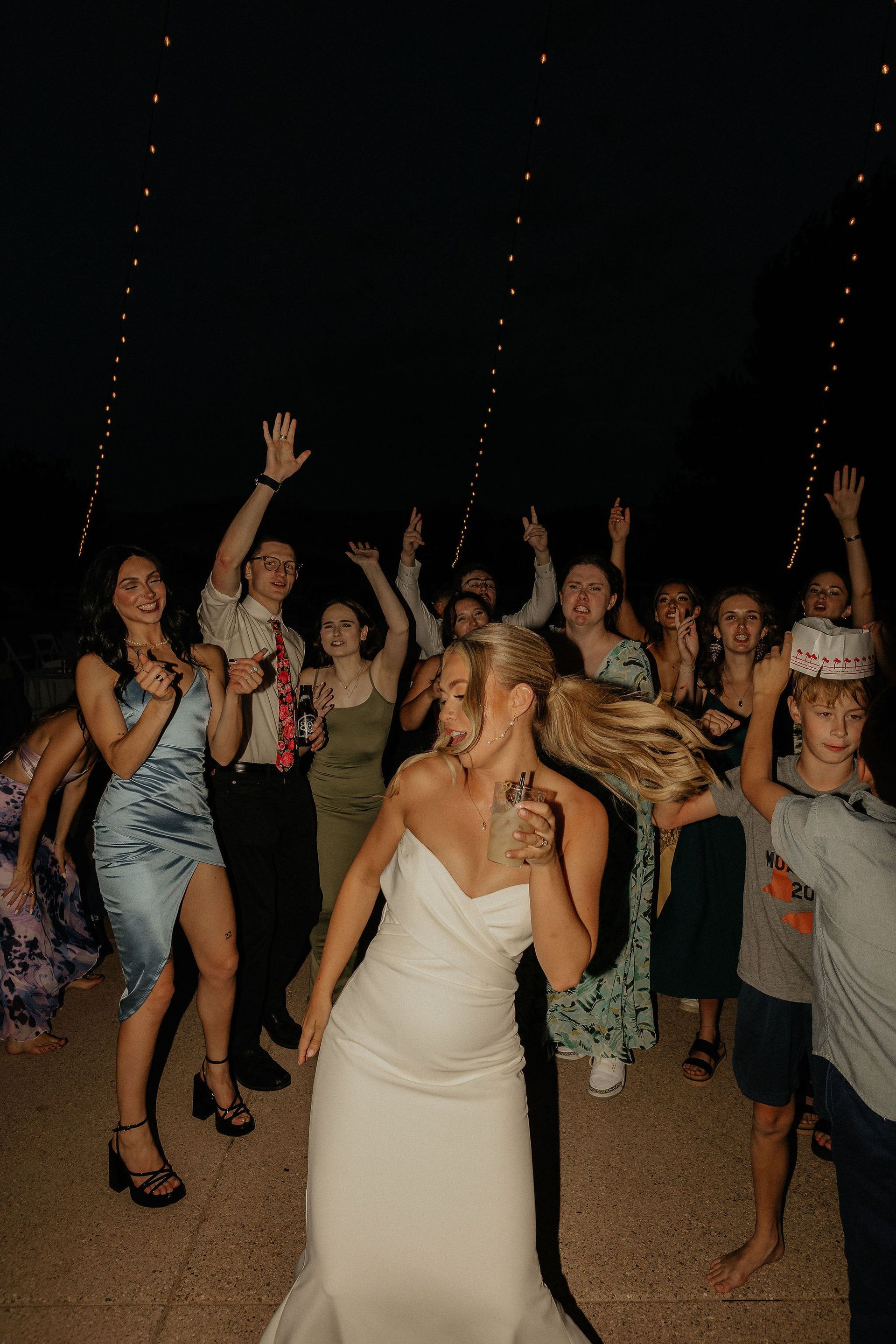 Bride in white dress celebrates with a cheering crowd at a nighttime outdoor party