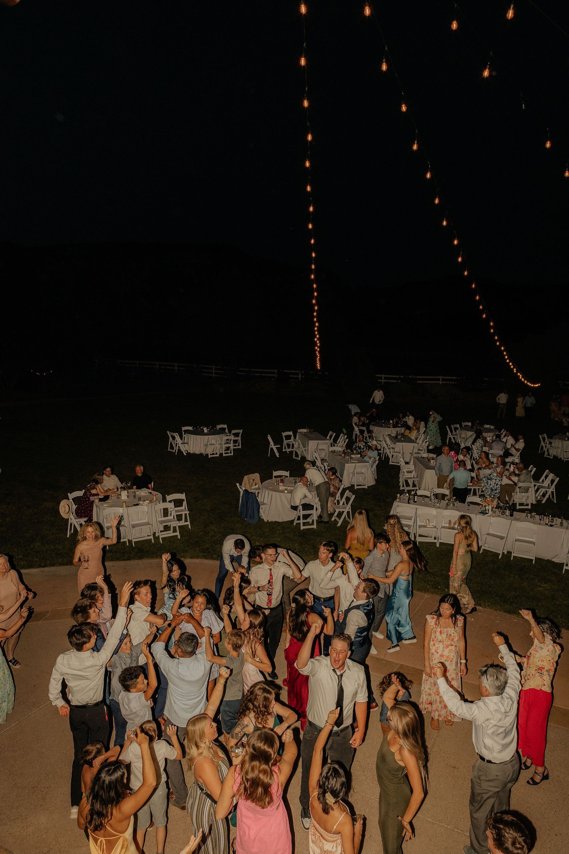 People dancing outdoors at night under string lights, with a crowd and tables in the background.