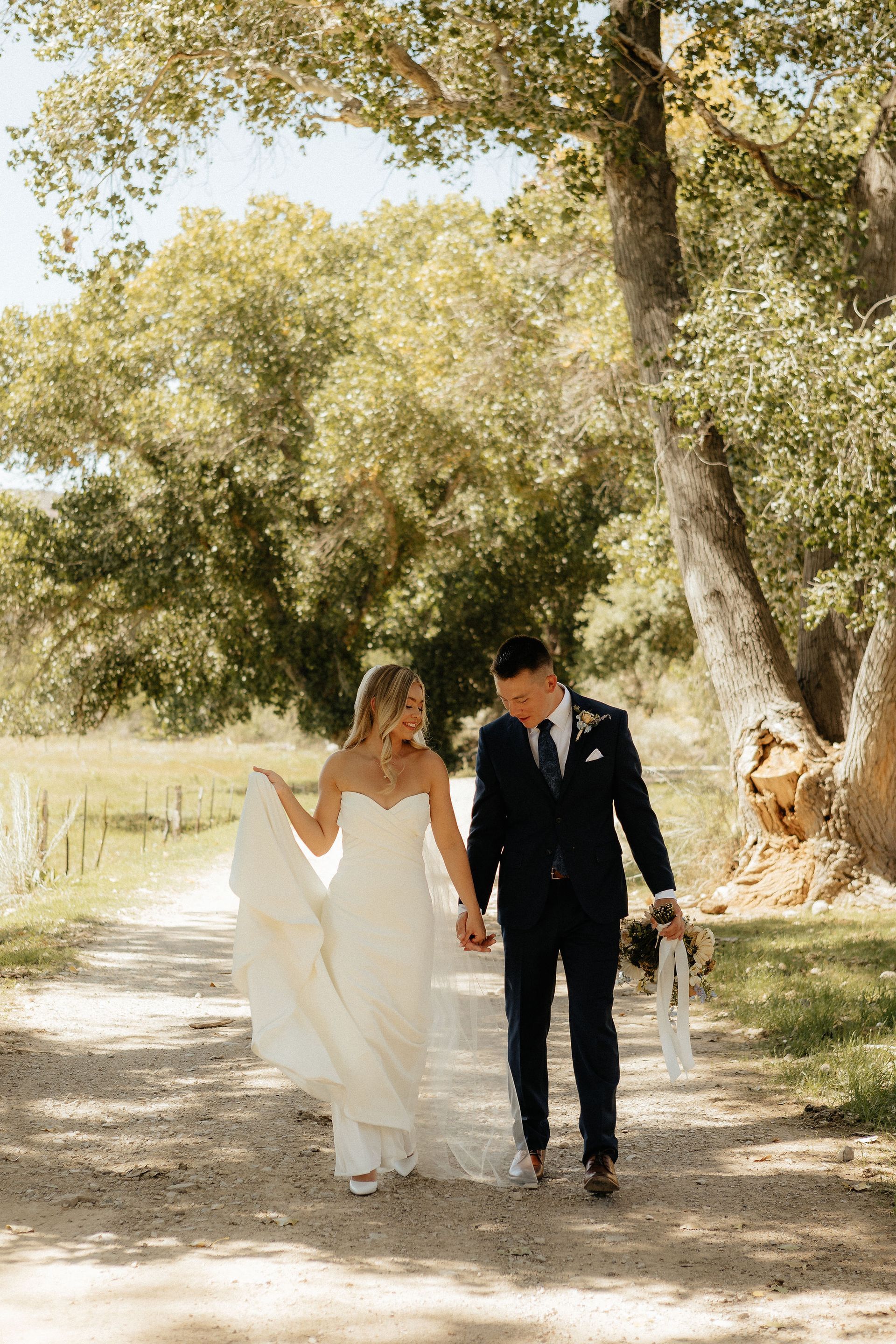 Bride and groom walking hand in hand under trees on a sunlit path